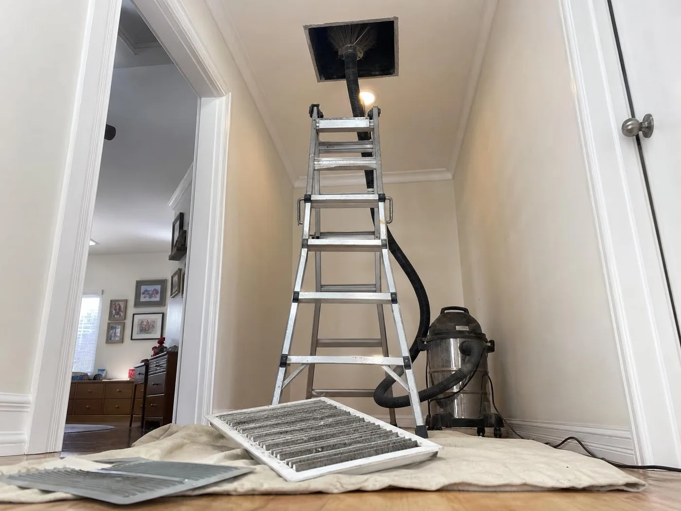 Hallway ceiling with the supply register removed and a rotating brush hose in the duct opening, ladder, shop vacuum and dusty registers on a drop cloth (illustration)