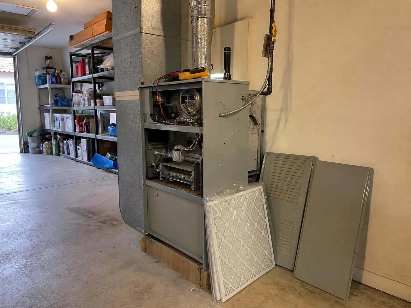Gas furnace in a garage with the access panels removed, burners and gas valve exposed, multimeter on top and a new filter leaning against it (illustration)