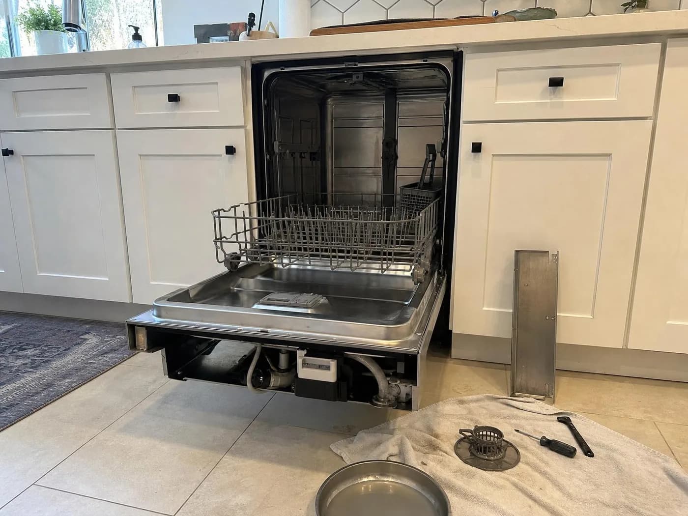 Built-in dishwasher pulled out of the cabinet during a repair, door open, filter and screwdriver on a towel on the floor (illustration)