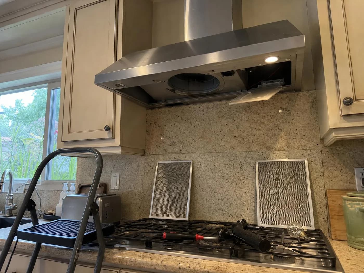 Wall-mounted range hood with both grease filters taken out and a light cover open, step stool and tools on the cooktop (illustration)